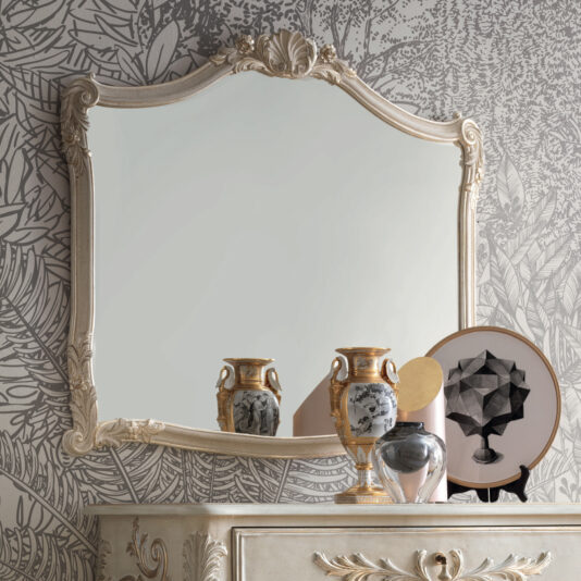 A vintage, ornate Classic Rococo Style Overmantel Mirror with a cream-colored frame hangs on a wall adorned with leafy black-and-white wallpaper. Below, a dresser showcases decorative items such as a round abstract art piece, ceramic vases, and a silver figurine.