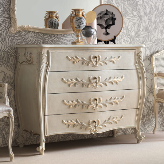 The Classic Style Bombe Chest Of Drawers, with its vintage charm and ornate design, features four elegant handles and intricate leaf detailing. Positioned against a floral wallpaper, it is adorned with decorative vases and a framed piece of art on the top surface, situated between two elegant chairs.