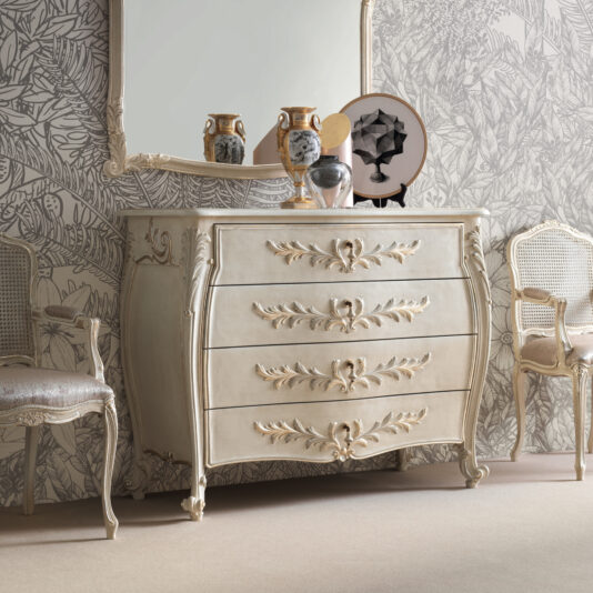 Classic Style Bombe Chest Of Drawers The elegant room showcases a Classic Style Bombe Chest Of Drawers, adorned with four intricately carved, gold-accented details. Above it hangs a large framed mirror, flanked by two ceramic vases and additional decor. Two upholstered chairs with matching ornate designs are placed on either side.