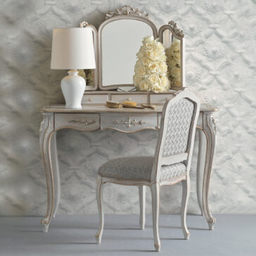 The Classic Style Dressing Table Set exudes a vintage charm with its ornate detailing, featuring a mirror, a white lamp, a vase of white roses, and gold-toned hairbrushes. Complementing this elegant vanity table is a matching chair adorned with a patterned cushioned seat, all set against textured floral wallpaper.