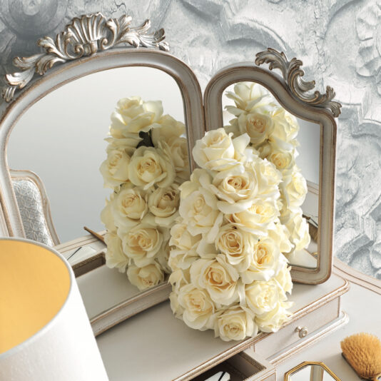 Classic Style Dressing Table Set A decorative arrangement of white roses is placed on the Classic Style Dressing Table Set, complemented by a vintage mirrored vanity. The mirrors reflect the bouquet, enhancing the lush, elegant look. The vanity features timeless details and is adorned with a nearby lamp and makeup brush. The background showcases patterned wallpaper, completing the sophisticated atmosphere.