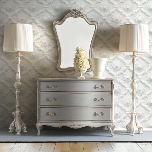 Classic Style Overmantel Mirror A vintage-style dresser with ornate handles sits against a floral-patterned wallpaper. On top, a decorative Classic Style Overmantel Mirror is framed by a vase of roses. Two elegant, tall lamps with pleated shades stand on either side of the dresser on a wooden floor, epitomizing classic style.