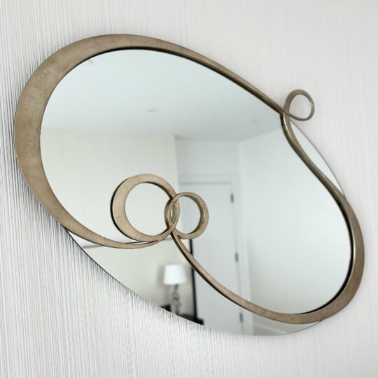 A Contemporary Twisted Oval Mirror, featuring an artistic and abstract metal frame, is mounted on a plain white wall. In its reflection, you can see part of a room with a closed white door, a white wall, and a table lamp with a white shade.