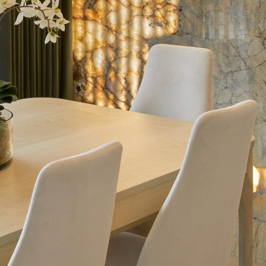 High-Back Curved Dining Chair A light-colored wooden dining table with four High-Back Curved Dining Chairs is set against a backdrop of marble walls and green curtains. A potted plant with white flowers is placed on the table, and warm sunlight illuminates the scene.
