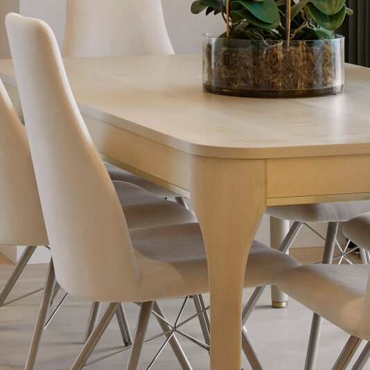 High-Back Curved Dining Chair A close-up of a modern dining room showcases a light wooden table paired with the High-Back Curved Dining Chairs, which are upholstered in white and feature metallic legs. Atop the table sits a transparent glass vase filled with green plants and moss. The background reveals a white wall and offers a glimpse of green curtains.