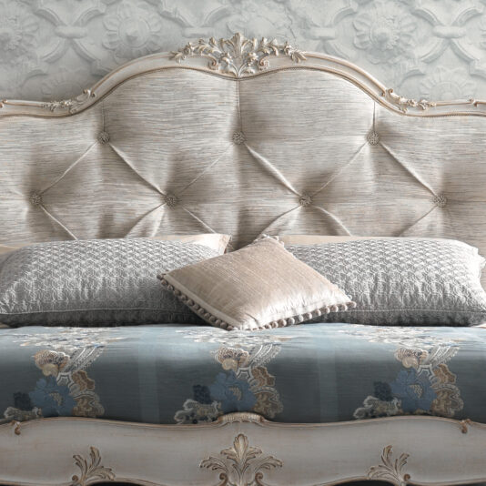 A close-up of the High End Button Upholstered Bed, elegantly showcased in a luxurious bedroom. The headboard is covered in soft, textured fabric and features intricate carvings and details. The bed is adorned with decorative pillows and a richly patterned bedspread.