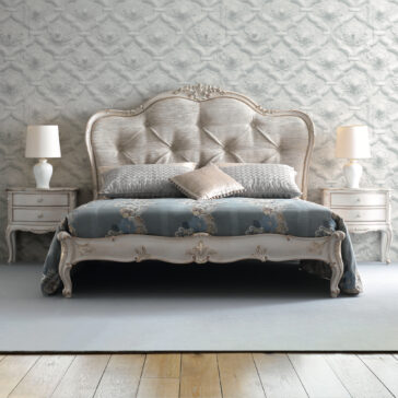An elegant bedroom showcases the High End Button Upholstered Bed, featuring a plush, tufted headboard and ornate wooden frame. The bed is adorned with blue and grey bedding and is flanked by two matching nightstands with white lamps. The backdrop is a textured, patterned wallpaper.