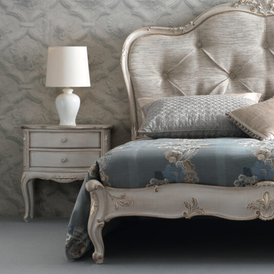 A bedroom designed with a retro charm featuring the High End Button Upholstered Bed, adorned with a blue-patterned bedspread and decorative pillows. Next to the bed, an antique wooden nightstand with two drawers is crowned by a white table lamp. The wall features an intricate, floral textured pattern.