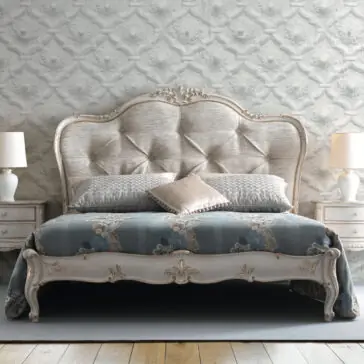 A bedroom featuring the High End Button Upholstered Bed, elegantly adorned with multiple pillows and a floral-patterned bedspread. The space includes matching bedside tables on both sides, each topped with a white lamp, set against a wall decorated with an ornate sculpted pattern in the background.