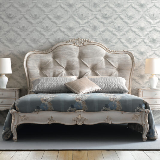 A bedroom featuring the High End Button Upholstered Bed, elegantly adorned with multiple pillows and a floral-patterned bedspread. The space includes matching bedside tables on both sides, each topped with a white lamp, set against a wall decorated with an ornate sculpted pattern in the background.