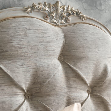 A close-up of the High End Button Upholstered Bed showcases its elegantly designed headboard, featuring a soft, textured fabric and tufted buttons that add a luxurious touch. The top edge is adorned with detailed floral carvings in a distressed antique finish, giving it a vintage and sophisticated appearance.