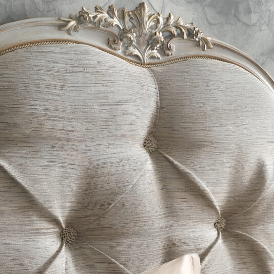 A close-up of the High End Button Upholstered Bed showcases its elegantly designed headboard, featuring a soft, textured fabric and tufted buttons that add a luxurious touch. The top edge is adorned with detailed floral carvings in a distressed antique finish, giving it a vintage and sophisticated appearance.