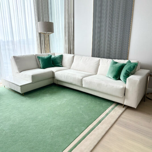 High End Corner Sofa Bed With Side Table A modern living room is showcased, featuring a High End Corner Sofa Bed With Side Table complemented by green accent pillows. The room includes a green carpet and a floor lamp with a beige shade near the large windows adorned with sheer white curtains. The walls are decorated with vertical textured panels.