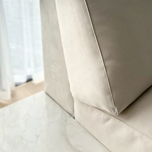 High End Corner Sofa Bed With Side Table A close-up of the High End Corner Sofa Bed With Side Table reveals neatly stitched cushions and a smooth, light beige suede texture. The image also includes part of a white marble coffee table and sheer curtains in the background, suggesting a bright, airy room.