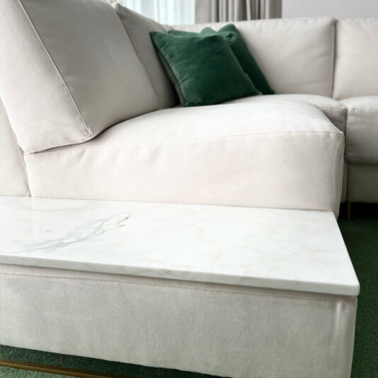 High End Corner Sofa Bed With Side Table A close-up of the High End Corner Sofa Bed With Side Table showcasing its modern white sectional design is featured. The smooth fabric upholstery harmonizes with a marble-topped coffee table attached to the sofa's base. Green accent pillows are neatly arranged in the corner, and green carpeting is visible below.