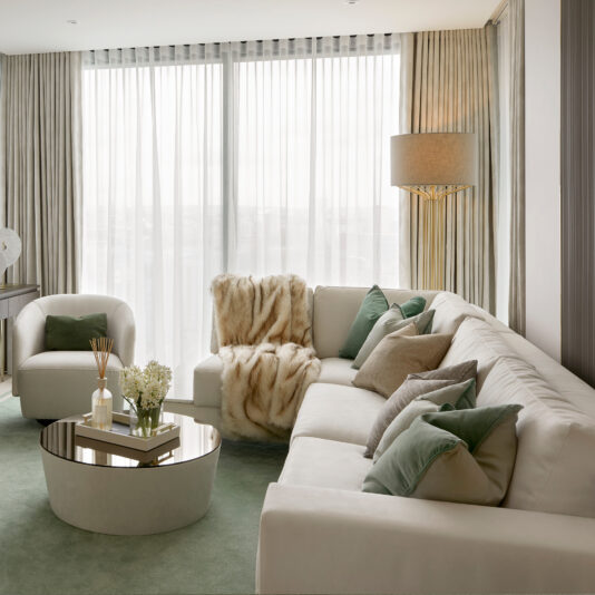 High End Corner Sofa Bed With Side Table A modern living room showcases a High End Corner Sofa Bed With Side Table, adorned with green and beige cushions and a fur throw. The space is complete with a round glass coffee table decorated with flowers and books, an armchair, and floor-to-ceiling windows with sheer curtains that allow natural light to flood the room.