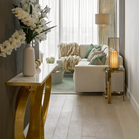 High End Corner Sofa Bed With Side Table Elegantly decorated living room featuring a High End Corner Sofa Bed With Side Table, adorned with green and beige cushions and complemented by a luxurious faux fur throw. A marble-topped console table with a large vase of flowers takes center stage in the foreground. Warm lighting, sheer curtains, and the chic gold side table enhance the sophisticated ambiance.