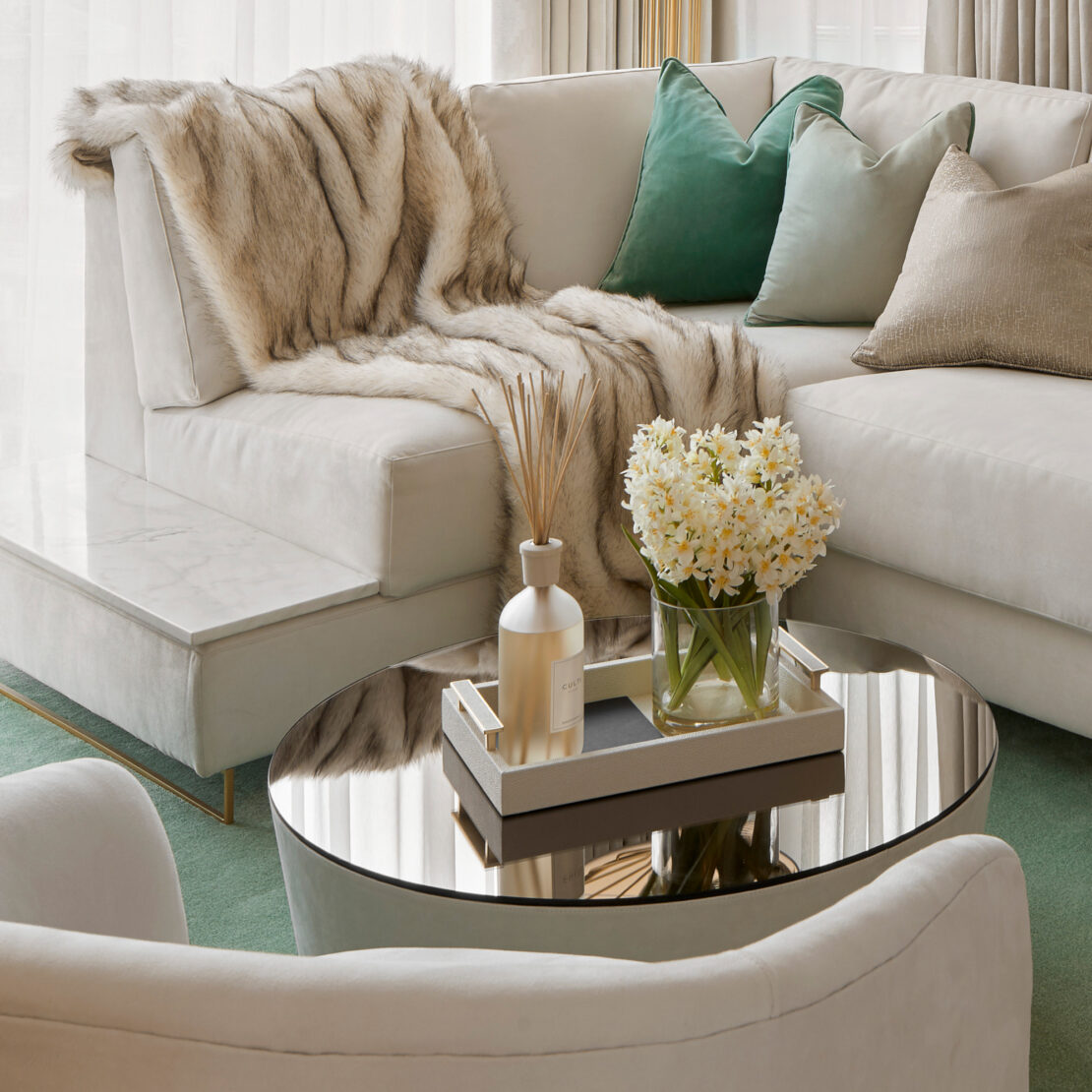 A modern living room showcases a high-end corner sofa bed with an integrated side table, adorned with a faux fur throw and green and beige pillows. A circular coffee table with a mirrored surface holds a tray of decorative items and a vase of white flowers. Beige curtains, along with an elegant additional side table and green rug, complete the space.