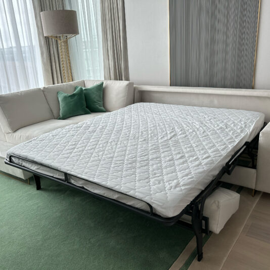 High End Corner Sofa Bed With Side Table A well-lit room features a white sofa with green accent pillows and a High End Corner Sofa Bed With Side Table. The sofa includes a pull-out bed, which is fully extended with a neatly made quilted mattress. The room has large windows with sheer curtains and a green area rug.