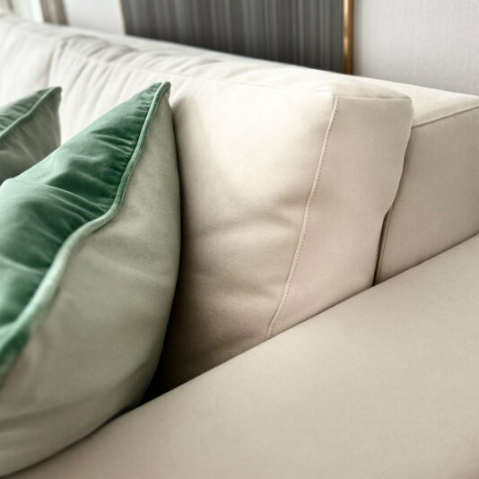 High End Corner Sofa Bed With Side Table Detailed view of the High-End Corner Sofa Bed with Side Table, showcasing two luxurious cushions. One cushion is light green trimmed with dark green velvet, while the other perfectly complements the sofa's light beige color. The integrated side table sits conveniently beside it, all set against a backdrop of subtle vertical paneling.