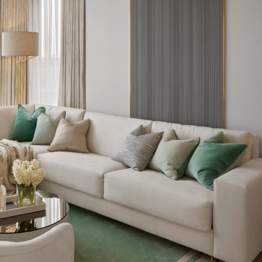 High End Corner Sofa Bed With Side Table A modern living room featuring a High End Corner Sofa Bed With Side Table adorned with various green and gray throw pillows. A glass coffee table with a bouquet of white flowers sits in front of the sofa. Neutral-toned curtains and a tall floor lamp are visible in the background, adding to the elegant ambiance.