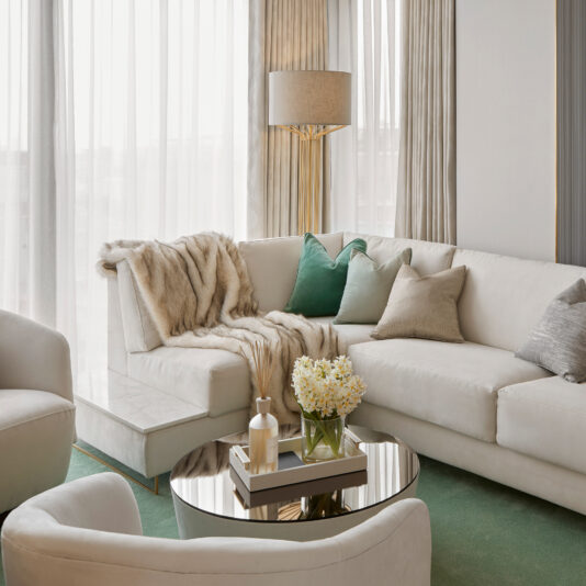 High End Corner Sofa Bed With Side Table A modern living room with a high-end corner sofa bed from our collection, complemented by green, beige, and gray pillows, and a fur throw. A glass coffee table stands nearby holding a vase of flowers and books. The space is illuminated by a tall floor lamp and features sheer white curtains, green carpeting, and an elegant side table from the high-end corner sofa bed set.