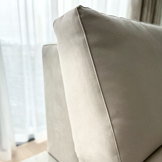 High End Corner Sofa Bed With Side Table Close-up of the seam on the backrest of a light gray suede **High End Corner Sofa Bed With Side Table**. Sheer white curtains softly diffuse light in the background, capturing the texture of the fabric and precise stitching detail on the cushion.