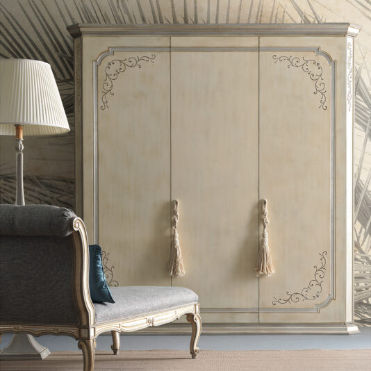 In a room with a patterned wall stands the *Large Classic Style Wardrobe*, featuring intricate, ornate carvings on its edges. This vintage wooden wardrobe boasts three doors, each adorned with a beige tassel handle. To the left is an elegant upholstered chaise lounge and a floor lamp.