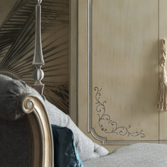 Large Classic Style Wardrobe Close-up of an elegant room featuring a grey upholstered chair with a wooden carved armrest, partially showing a blue cushion. In the background is a detailed wall panel with intricate carvings, a beige tassel hanging by its side, and the edge of the Large Classic Style Wardrobe.