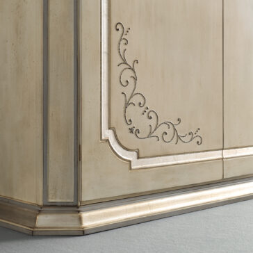 Large Classic Style Wardrobe Close-up of the Large Classic Style Wardrobe corner reveals its ornate, antique wooden design. It features intricate, curving carved patterns on its light beige surface with a beautifully weathered finish. The wardrobe is adorned with gold accents along its edges and rests on a matching baseboard against a gray floor.