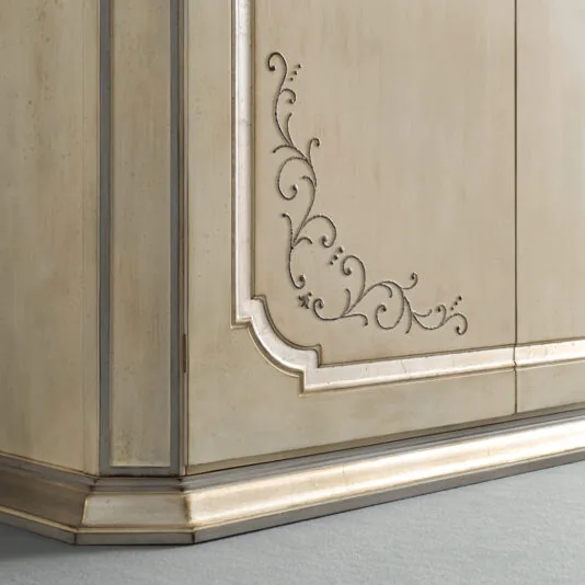Large Classic Style Wardrobe Close-up of the Large Classic Style Wardrobe corner reveals its ornate, antique wooden design. It features intricate, curving carved patterns on its light beige surface with a beautifully weathered finish. The wardrobe is adorned with gold accents along its edges and rests on a matching baseboard against a gray floor.