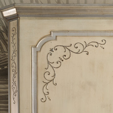 Large Classic Style Wardrobe A close-up of the Large Classic Style Wardrobe reveals a cream-colored wooden cabinet with intricate, carved floral designs on its door. The edges feature ornate detailing highlighted in a silver finish. The background shows a faint sketch-like pattern.