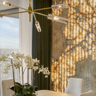 A modern dining room features a Mid-Century Modern Chandelier with tubular lights, an orchid centerpiece on the table, white upholstered chairs, and a large textured backlit marble wall. The space is enhanced by floor-to-ceiling windows with sheer and dark green curtains.
