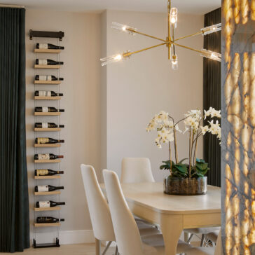 A contemporary dining room showcases a light-colored wooden rectangular table and white cushioned chairs. The space is illuminated by a Mid-Century Modern Chandelier with exposed bulbs that hangs above. Additional elements include a vertical wine rack, black curtains, and a pot of white orchids placed at the table's center.