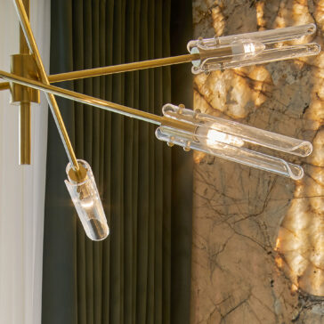 The **Mid-Century Modern Chandelier** featuring transparent glass bulbs and gold metal fixtures is mounted on the ceiling. It is positioned near dark green curtains and a marble wall with intricate veining, softly illuminated by the light from its bulbs.