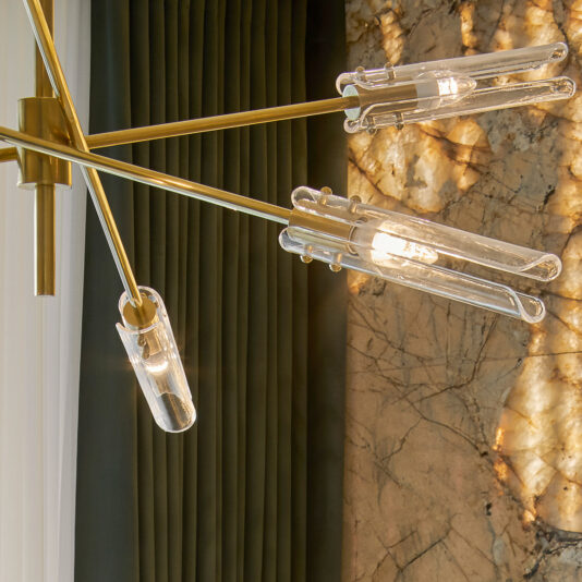 Mid-Century Modern Chandelier The **Mid-Century Modern Chandelier** featuring transparent glass bulbs and gold metal fixtures is mounted on the ceiling. It is positioned near dark green curtains and a marble wall with intricate veining, softly illuminated by the light from its bulbs.