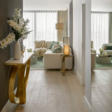 A modern, elegantly furnished living room with a white sofa, green accents, and a large window with sheer curtains. A hallway features the sleek Modern Narrow Marble Ring Console stylishly adorned with a vase of white flowers. Additionally, there's a tall lamp and a side table that holds another lamp in view.