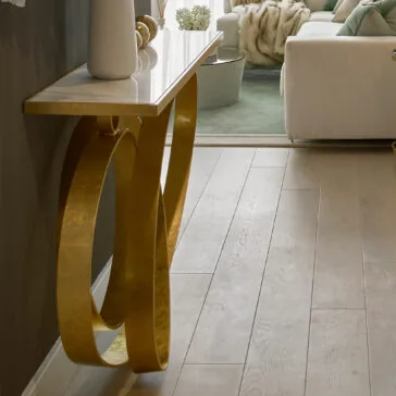 A modern living room features the **Modern Narrow Marble Ring Console** table, complete with a sophisticated white marble top and an elegant gold frame. The table is tastefully adorned with decorative items. A portion of a white sofa with a green throw blanket is visible, set against light wood flooring and complemented by a green area rug in the background.