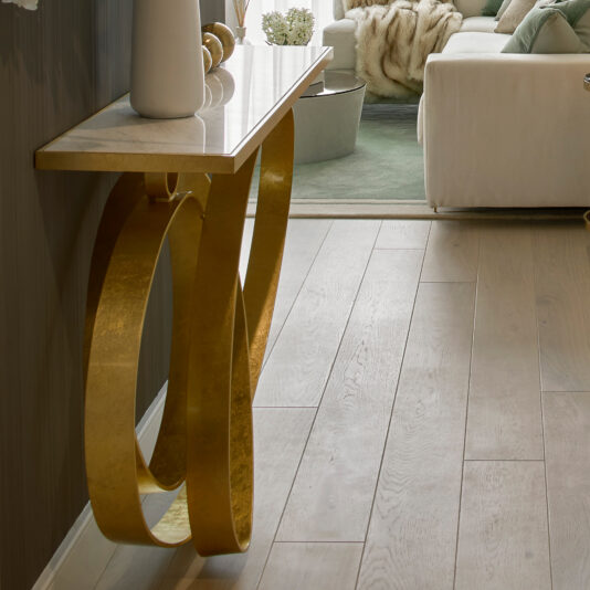 A modern living room features the **Modern Narrow Marble Ring Console** table, complete with a sophisticated white marble top and an elegant gold frame. The table is tastefully adorned with decorative items. A portion of a white sofa with a green throw blanket is visible, set against light wood flooring and complemented by a green area rug in the background.