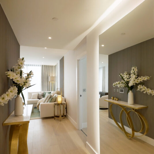 Modern Narrow Marble Ring Console A modern hallway features light wood floors, a large wall mirror, the Modern Narrow Marble Ring Console, white flower arrangements, and opens into a bright living room with a white sofa and neutral decor.