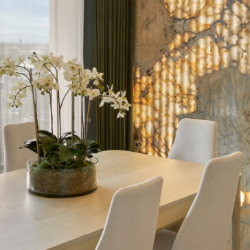 A dining area with the Modern White Maple Dining Table surrounded by white, high-backed chairs. A round glass vase with white orchid flowers sits on the table. The backdrop features a marble wall with intricate lighting, next to large green curtains and a bright window.