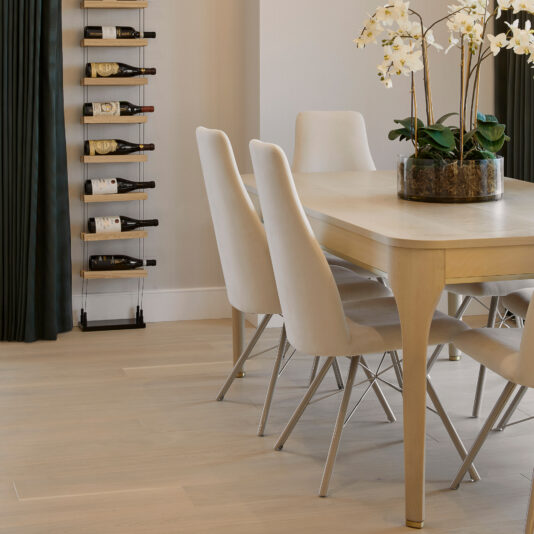 A modern dining room features a sleek Modern White Maple Dining Table accompanied by six white chairs with metal legs. The table holds a decorative plant adorned with white flowers. A minimalist wine rack, stocked with multiple bottles, is mounted on the wall near the dark drapes. Light wood flooring completes this elegant space.