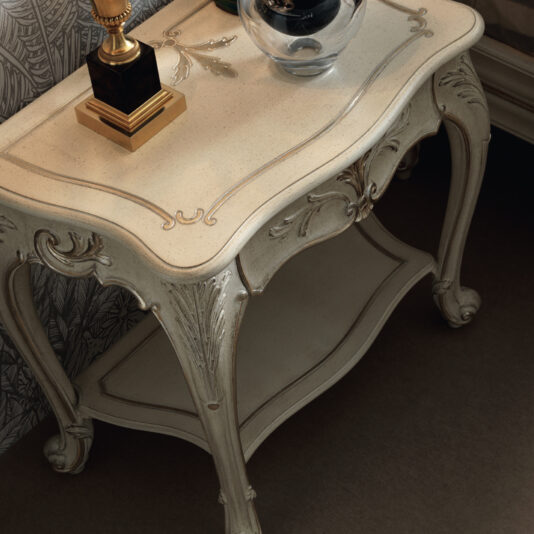 An Ornate Classic Style Bedside Table with intricate carvings and a distressed white finish graces the room. This vintage piece holds a brass lamp with a square base and a black decorative glass orb. The background showcases detailed wallpaper with a leaf pattern, and the table features an open lower shelf.