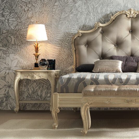 Ornate Classic Style Bedside Table An elegant bedroom showcases a bed with an ornate, tufted headboard in a neutral tone, patterned bedding, and an Ornate Classic Style Bedside Table adorned with intricate carvings. A modern table lamp emits a warm glow, set against detailed, nature-inspired wallpaper.