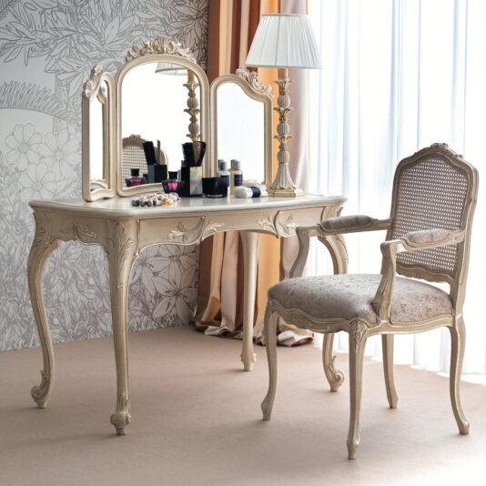 The Ornate Classic Style Dressing Table Set showcases elegant intricate detailing and a large mirror flanked by two side panels. The surface is adorned with various cosmetics, and placed in front is a vintage-style cushioned chair featuring a wicker backrest and armrests. Completing the setup, a lamp rests atop the table.