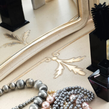 Ornate Classic Style Dressing Table Set A close-up of the Ornate Classic Style Dressing Table Set showcases its vanity table with an ornate mirror frame adorned with decorative leaf designs. This elegant dressing table holds various jewelry pieces, such as pearl and metallic bead necklaces, alongside makeup and perfume bottles in black and dark tones.