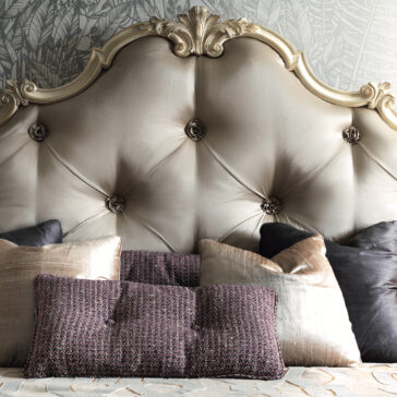 The Romantic Button Upholstered Bed, adorned with an elegant tufted headboard featuring elaborate detailing and six buttons, stands out beautifully against a patterned wallpaper. The bed is tastefully furnished with plush pillows in diverse textures and colors, including metallic and dark shades, bringing a touch of luxury to the bedroom decor.