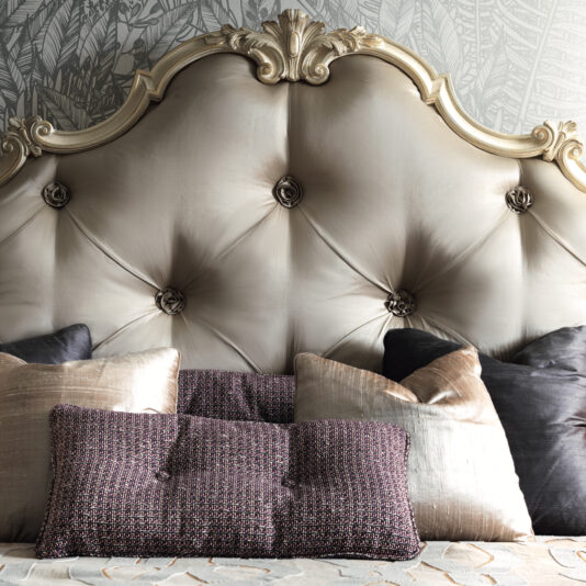 Romantic Button Upholstered Bed The Romantic Button Upholstered Bed, adorned with an elegant tufted headboard featuring elaborate detailing and six buttons, stands out beautifully against a patterned wallpaper. The bed is tastefully furnished with plush pillows in diverse textures and colors, including metallic and dark shades, bringing a touch of luxury to the bedroom decor.
