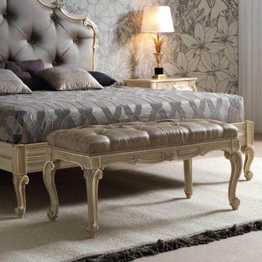A luxurious bedroom showcases an ornate bed with a tufted, upholstered headboard and a patterned bedspread. Positioned at the foot of the bed is a Small Louis Style Bench featuring elegantly curved legs. A lit bedside lamp on a nightstand adds warmth to the space while floral wallpaper graces the wall.