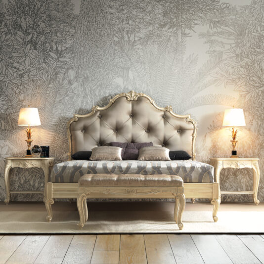 Luxurious bedroom with an ornate, tufted headboard in cream. Two elegant nightstands with matching carved wood details flank the bed, each topped with a lamp. The wall behind features intricate botanical wallpaper, and a graceful Small Louis Style Bench sits at the foot of the bed.